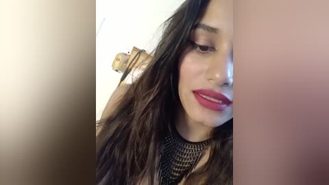 Media: Video of a young woman with long, wavy dark hair, fair skin, and red lipstick, wearing a black, textured top. Background is blurred, showing a beige wall and part of a wooden chair.