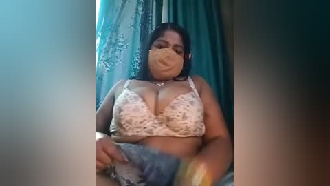 Media: Video of a plus-sized, dark-skinned woman with a large bust, wearing a floral bra and face mask, sitting indoors.