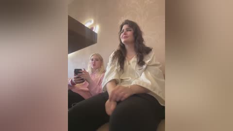 Media: Video of two women, one with long, dark hair, in a beige room with floral wallpaper. Both wear casual outfits; one holds a smartphone.