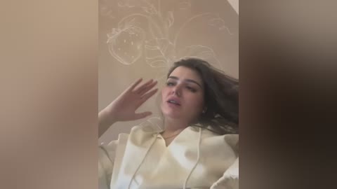 Media: A video captures a young woman with long dark hair and fair skin, wearing a white jacket, her hands raised, and eyes closed, set against a beige wall with faint floral designs.