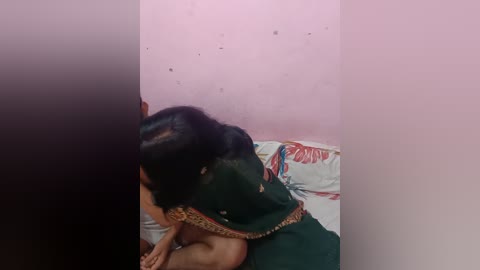 Media: A video shows a woman with dark hair, wearing a green sari, sitting on a bed with floral bedding, in a pink-walled room.