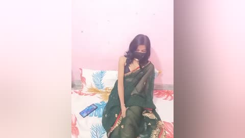 Media: Video of a South Asian woman in a dark green sari with gold embroidery, sitting on a bed with a floral-patterned sheet, wearing a black face mask, holding a phone. Pink walls, dim lighting.