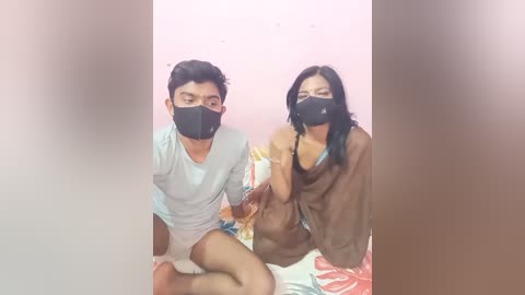 Media: A video of a man and woman, both wearing face masks, sitting on a bed in a dimly lit room with a pink wall. The man is in light clothing, while the woman wears a brown top.