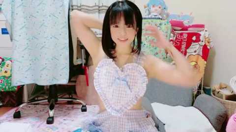 Media: Video of a young Asian woman with straight black hair, wearing a white and blue plaid dress with a heart cutout, sitting on a pink floor, surrounded by toys and clothes in a cluttered room.