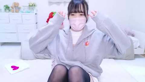 Media: A video of an Asian woman with a mask, wearing a gray hoodie, black tights, and a white top, sitting on a bed in a white room with a dresser, teddy bear, and a red Santa hat.