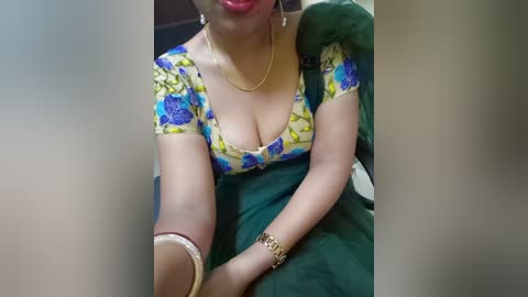 Media: Video of a woman with light brown skin, wearing a vibrant floral blouse and gold jewelry, reclining on a dark green bed.