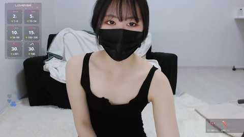 Media: Video of an East Asian woman with short black hair, wearing a black face mask, black tank top, and black pants, sitting on a white rug in a simple, cluttered room with a black sofa and a calendar.