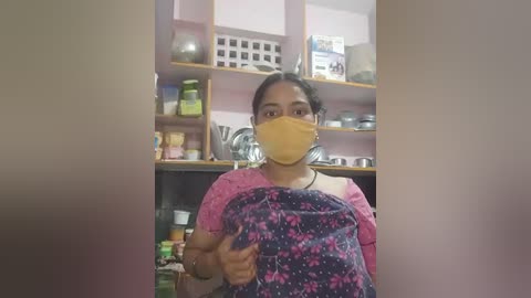 Media: Video of a South Asian woman with medium brown skin, wearing a yellow face mask and a pink floral-patterned sari, standing in a cluttered kitchen with pink cabinets and various household items.