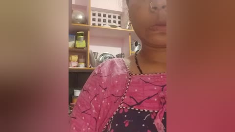 Media: Video of a young person with light skin, wearing a pink floral-patterned shirt, partially visible in a cluttered kitchen with a white and brown background.