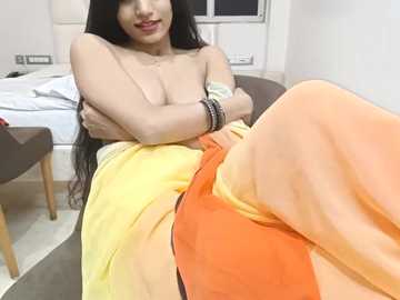 Media: Video of a topless woman with long black hair, medium breasts, and fair skin, sitting in a chair, draped in a yellow and orange sari, with a white bed and brown chair in the background.