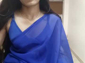 Media: Video of a woman with long black hair, wearing a sheer blue sari, partially revealing her medium-sized breasts and a delicate gold necklace. The background features a beige wall and a partially visible brown door.