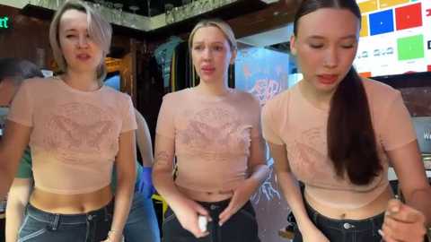 Media: Video of three young women with blonde hair, wearing matching pink graphic tees, dark jeans, and casual expressions in a dimly lit, colorful indoor setting.