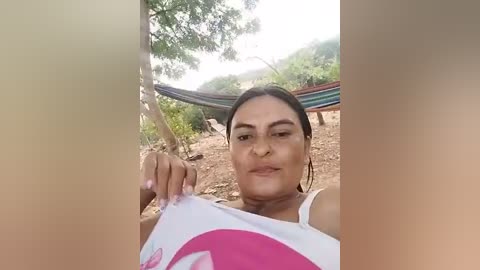 Media: A video of a Latina woman with medium skin tone, dark hair, and a slender build, wearing a white tank top with pink floral design. She sits outdoors on a dirt ground, near a hammock, under a tree.