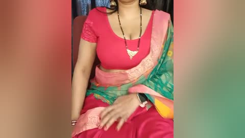 Media: Video of a South Asian woman in a red blouse and green sari with a pink border, seated, wearing gold earrings and a necklace.