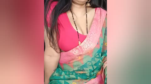 Media: Video of a South Asian woman with medium skin tone, wearing a vibrant pink blouse, a semi-transparent green and pink sari with intricate designs, and a black beaded necklace.