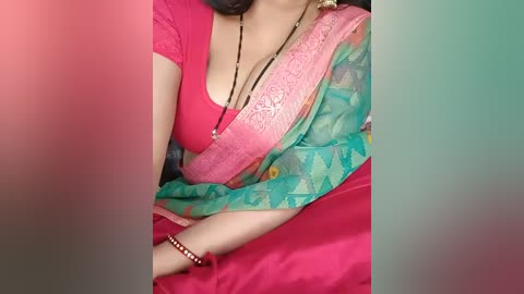 Media: Video of a woman with medium skin tone wearing a vibrant pink top and a green sari with a floral pattern. Her dark hair is partially visible, and she wears a black beaded necklace.