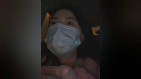 Media: Video of an Asian woman in a dimly lit room, wearing a blue surgical mask and a pink fluffy robe. Her face is partially obscured by shadows and a white object.