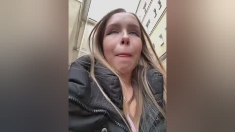 Media: Video of a Caucasian woman with long, straight blonde hair, fair skin, and a neutral expression, wearing a black puffer jacket. She stands in a narrow, indoor alleyway with beige and white buildings in the background.