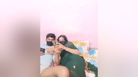 Media: Video of a couple in a bed, a man in a white shirt and a woman in a green sari, holding a phone, against a pink wall, with a blurry background.