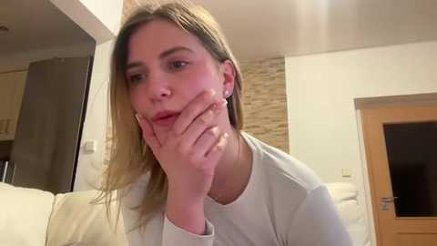 Media: Video of a Caucasian woman with straight, blonde hair, wearing a white shirt, resting her chin on her hand, in a modern, beige-toned living room with a stone accent wall and wooden door.