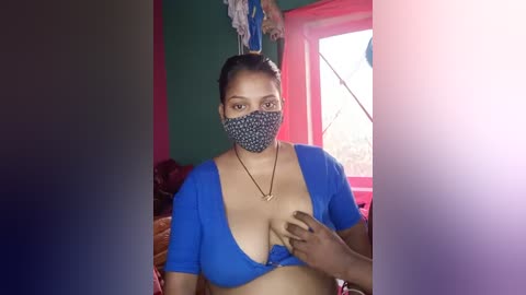 Media: Video of a woman with medium skin tone and medium-sized breasts, wearing a blue crop top and face mask, standing in a room with green walls, a red window, and a person hanging laundry.