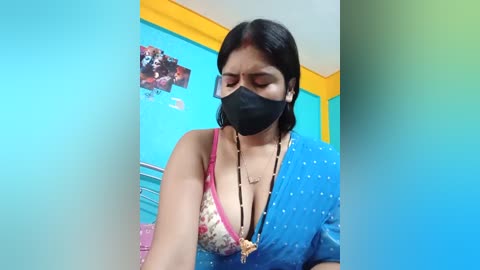 Media: Video of a South Asian woman with medium skin tone, wearing a black face mask, pink bra, and blue saree, standing in a brightly colored room.