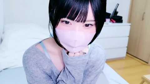 Media: A video of an East Asian woman with short black hair and pale skin, wearing a light pink face mask and gray long-sleeve shirt, kneeling in a modern, minimalist bedroom with white furniture and wooden floors.