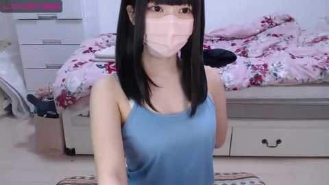 Media: A video of an Asian woman with straight black hair, wearing a blue tank top, face mask, and sitting on a pink floral bedspread in a bedroom with white furniture and a dresser.