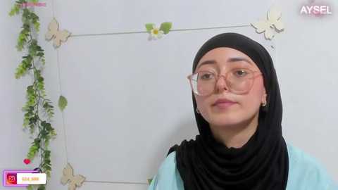 Media: Video of a young Middle Eastern woman with fair skin, wearing a black hijab and light blue top, sitting against a white wall adorned with butterfly decals and green vines.