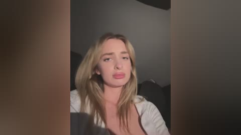 Media: Video of a young, fair-skinned, blonde woman with straight hair, wearing a white top, sitting in a dimly lit room. She has a neutral expression.