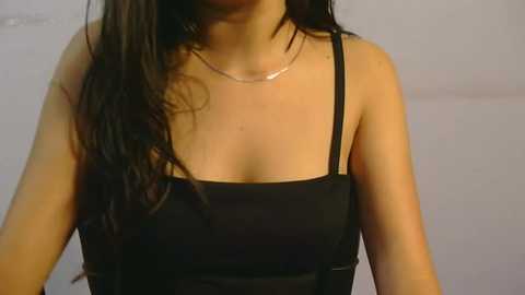 Media: Video of a woman with long black hair, wearing a black spaghetti-strap top and a thin silver necklace, standing against a plain, light gray background.