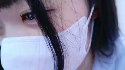 Media: Video of an East Asian woman with long, straight black hair, wearing a white surgical mask, covering most of her face except for her eyes, which are dark and expressive. She has a soft, pale complexion and is dressed in a light blue top.