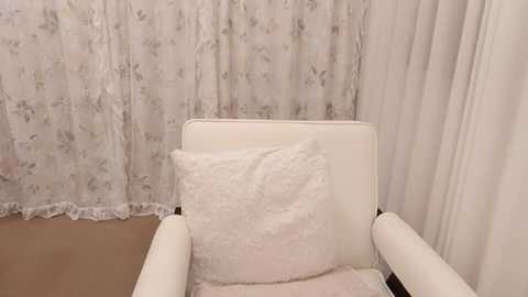 Media: Video of a cozy, white upholstered armchair with a fluffy white cushion, set against a backdrop of sheer white curtains with floral patterns. The room's floor is carpeted in a light beige color.