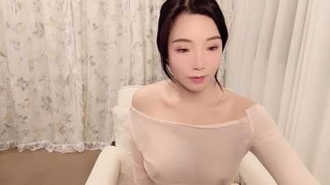 Media: A video of an Asian woman with fair skin and long, dark hair, wearing a sheer, off-the-shoulder top, seated in a white chair against floral-patterned curtains in a soft, beige room.