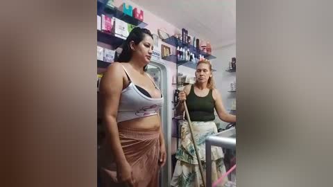 Media: A video of two women, one wearing a gray crop top and pink skirt, the other in a green tank top and floral skirt, standing in a cluttered kitchen.