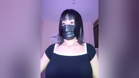 Media: Video of an East Asian woman with shoulder-length black hair and bangs, wearing a black face mask, black top, and white choker, standing indoors in a dimly lit room with white walls and a door.