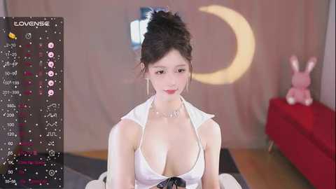 Media: Video of an Asian woman with fair skin, dark hair in a bun, wearing a revealing white halter top, sitting indoors near a red couch and a stuffed bunny.