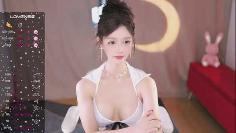 Media: Photorealistic CGI of a young East Asian woman with dark hair in a messy bun, wearing a revealing white top, sitting in a modern room with a pink bunny toy and a red sofa.