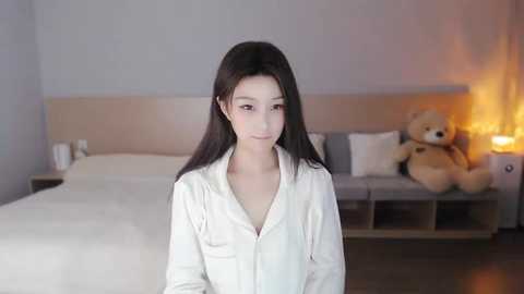 Media: Video of a young Asian woman with long black hair, wearing a white button-down shirt, standing in a modern, dimly lit bedroom with a beige headboard, a plush teddy bear, and a nightstand with a lamp.