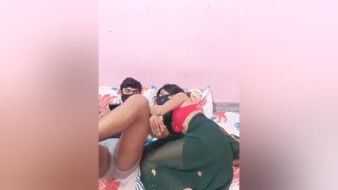 Media: Video of two young women with medium brown skin, lying on a bed with floral sheets. One wears a red top and black shorts, the other a green sari. They appear to be in a playful, intimate pose.