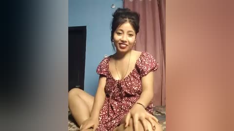 Media: Video of a smiling, dark-skinned Asian woman with medium-length black hair in a bun, wearing a red floral dress, sitting on a patterned carpet in a room with blue and pink walls.