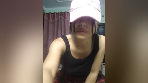 Media: Video of a woman in a black tank top, wearing a white baseball cap, with a blurred background featuring pink curtains and a dark wooden shelf.