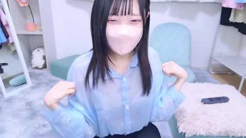 Media: Video of an Asian woman with straight black hair, wearing a light blue sheer blouse and a face mask, sitting on a light blue chair in a cozy, pastel-colored room with a fluffy rug.