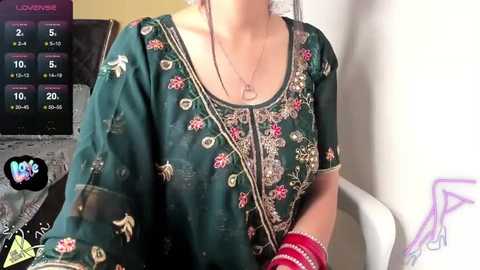 Media: Video of a fair-skinned woman in a green floral embroidered saree, wearing red bangles and a necklace, seated indoors with a medical monitor in the background.