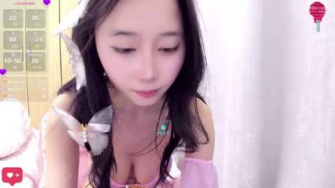 Media: Video of an East Asian woman with long black hair and fair skin, wearing a pink off-shoulder top, looking down, in a bedroom with white curtains.