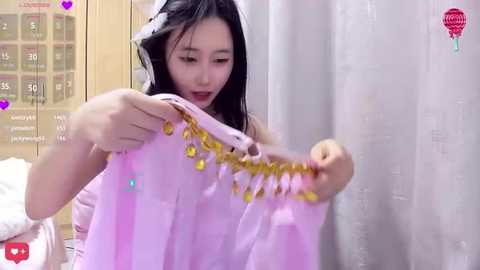 Media: A young Asian woman with long black hair, wearing a pink dress, examines a string of golden beads. The background features a white curtain and a wooden cabinet.