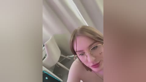 Media: A video of a Caucasian woman with fair skin and light brown hair, wearing glasses and red lipstick, bending over a white chair in a dimly lit room with white curtains.