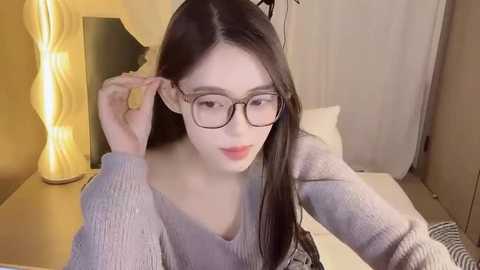 Media: Video of a young Asian woman with long black hair, fair skin, and large rectangular glasses, adjusting her glasses in a cozy bedroom with a beige lamp and a white curtain.