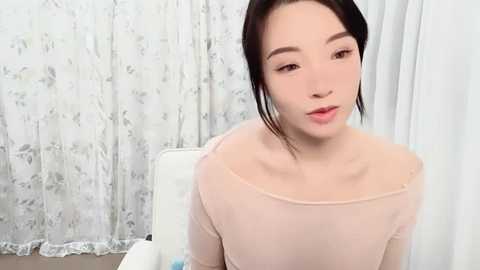 Media: Video of a young East Asian woman with light skin and straight black hair, wearing a sheer, off-the-shoulder top, seated in a white chair with floral-patterned curtains in the background.