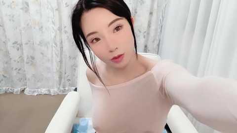 Media: A video of an East Asian woman with fair skin, dark hair, and a round face, wearing a light pink long-sleeve top, sitting in a white bathtub, with white floral-patterned curtains in the background.